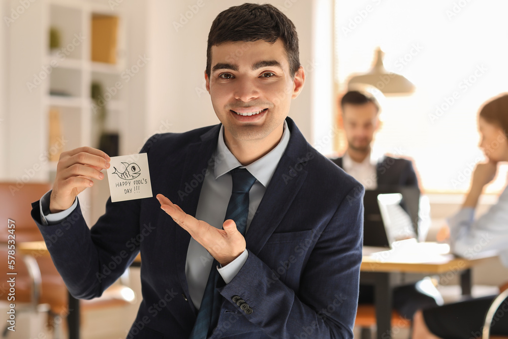 Young man showing sticky paper with text HAPPY FOOLS DAY in office