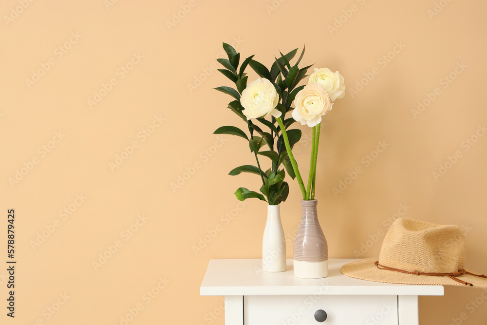 Vases with ranunculus flowers, plant branches and hat on table near beige wall