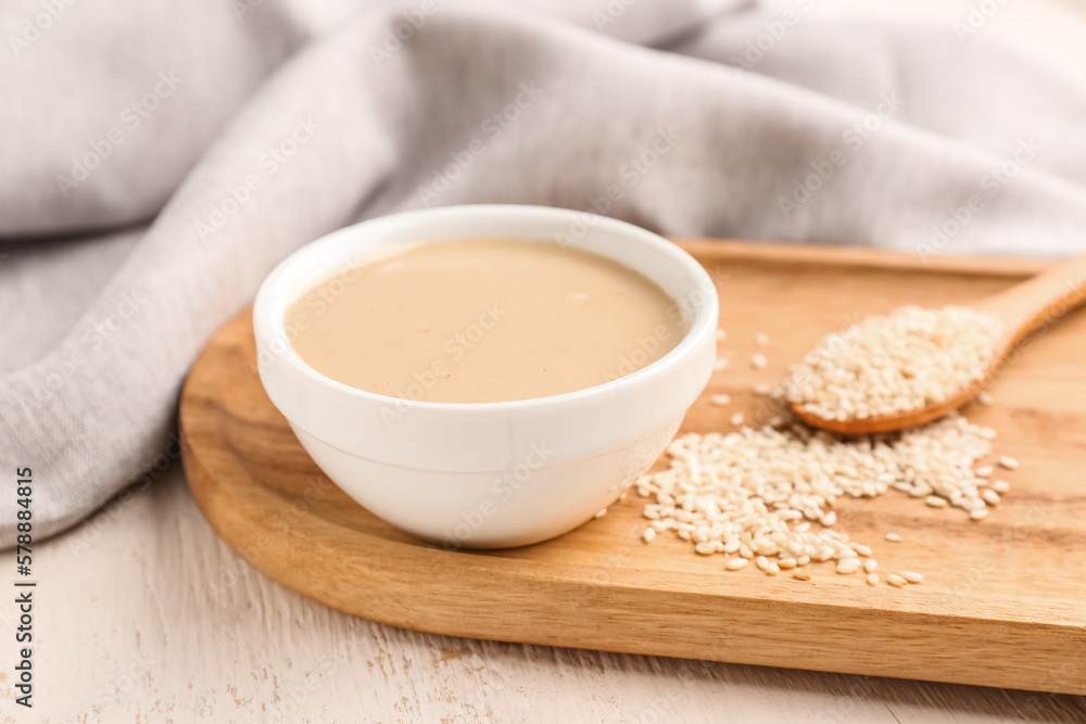 Board with bowl of tasty tahini and sesame seeds on light wooden table, closeup