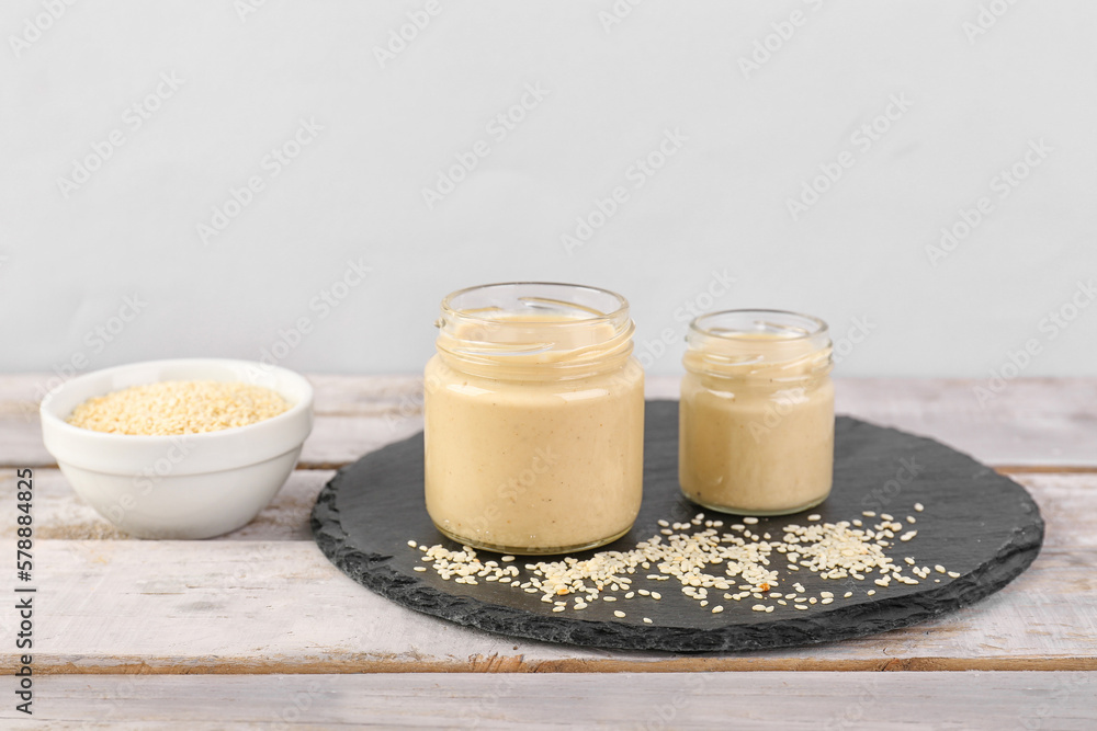 Slate board with jars of tasty tahini and sesame seeds on light wooden table