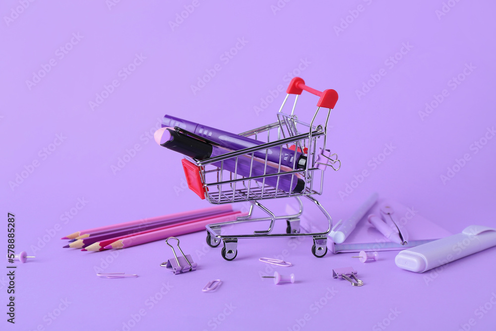 Shopping cart with different stationery on lilac background