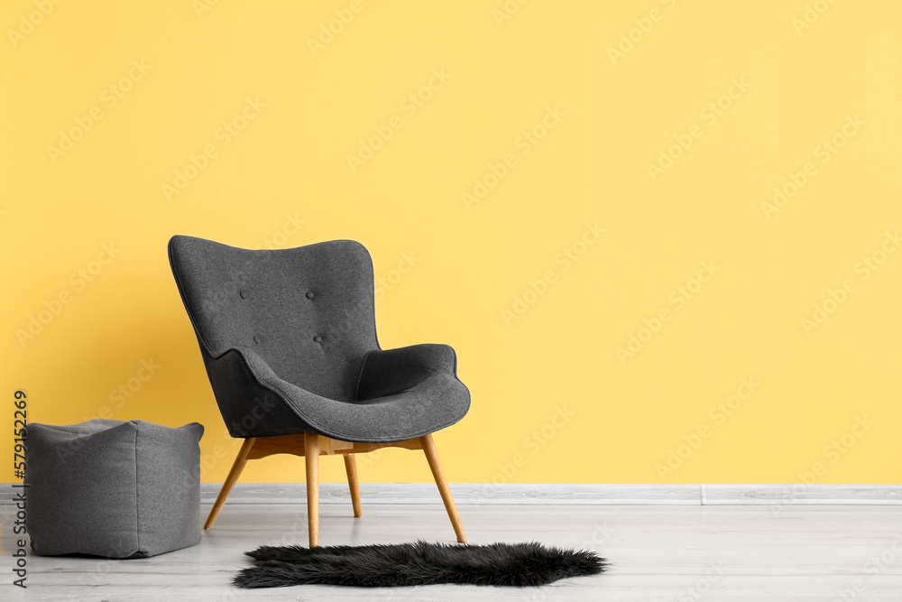 Stylish grey armchair, pouf and black fur rug near yellow wall