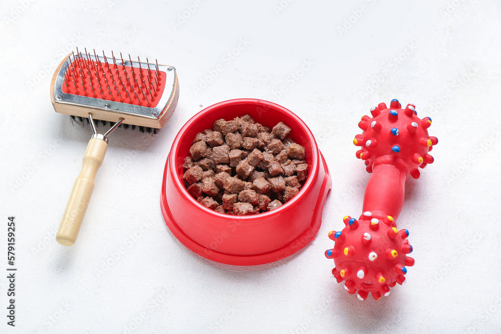 Bowl of wet pet food, toy and grooming brush on light background