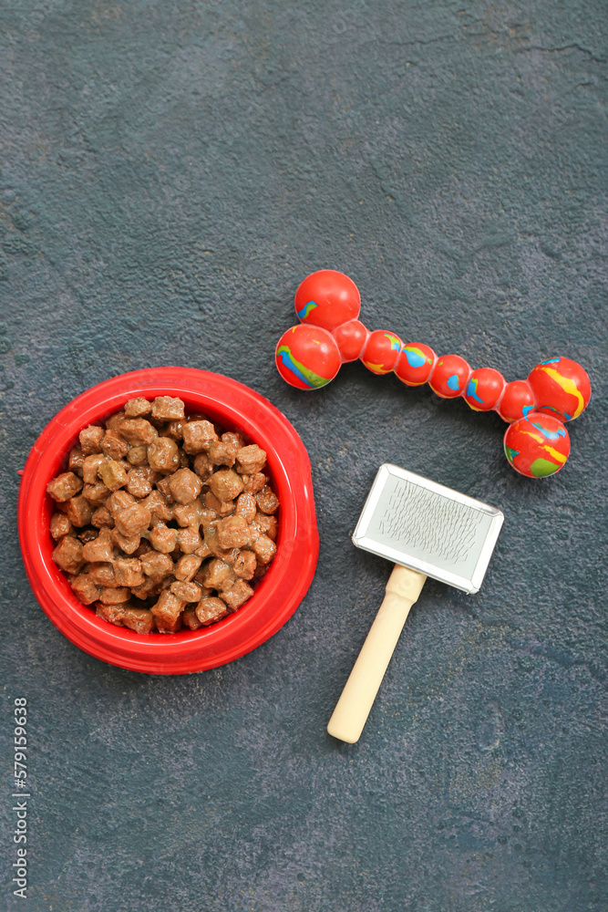 Bowl of wet pet food, rubber toy and grooming brush on dark background