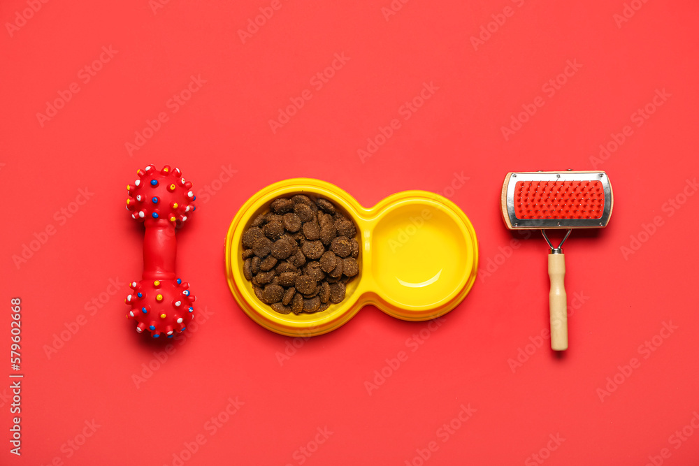 Bowl of dry pet food, grooming brush and rubber toy on red background