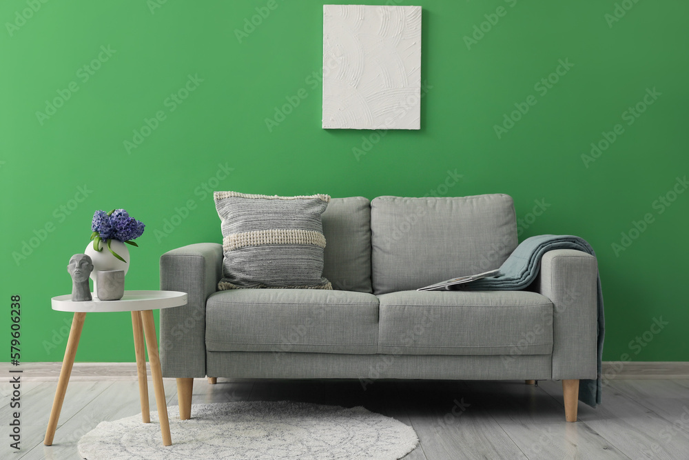 Grey sofa with cushion, plaid and vase of hyacinth flowers on table near green wall