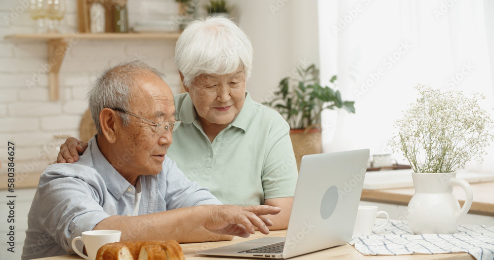 Happy asian senior couple sitting at kitchen table surfing web on laptop together, doing online shop