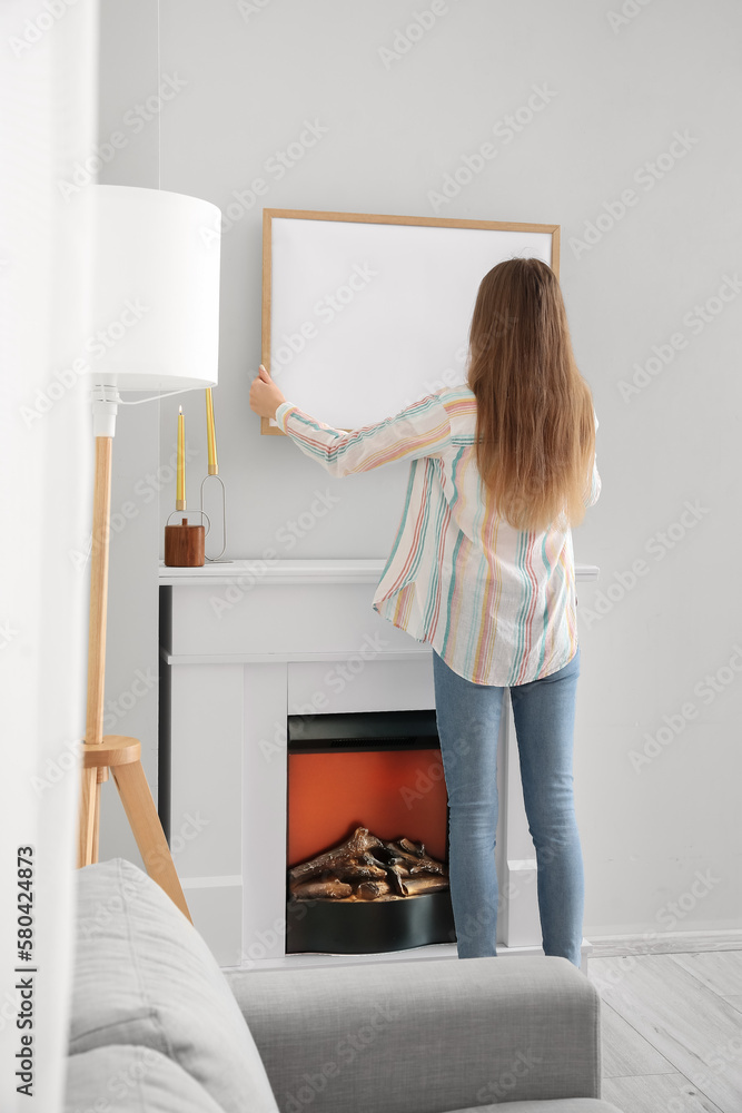 Young woman hanging blank frame on light wall at home, back view