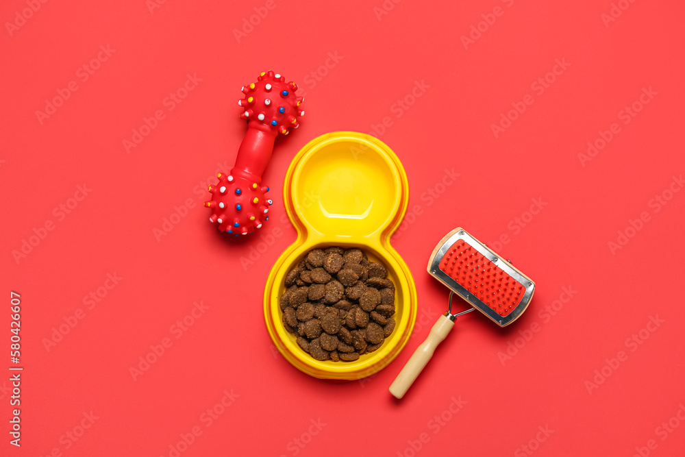 Bowl of dry pet food, grooming brush and rubber toy on red background