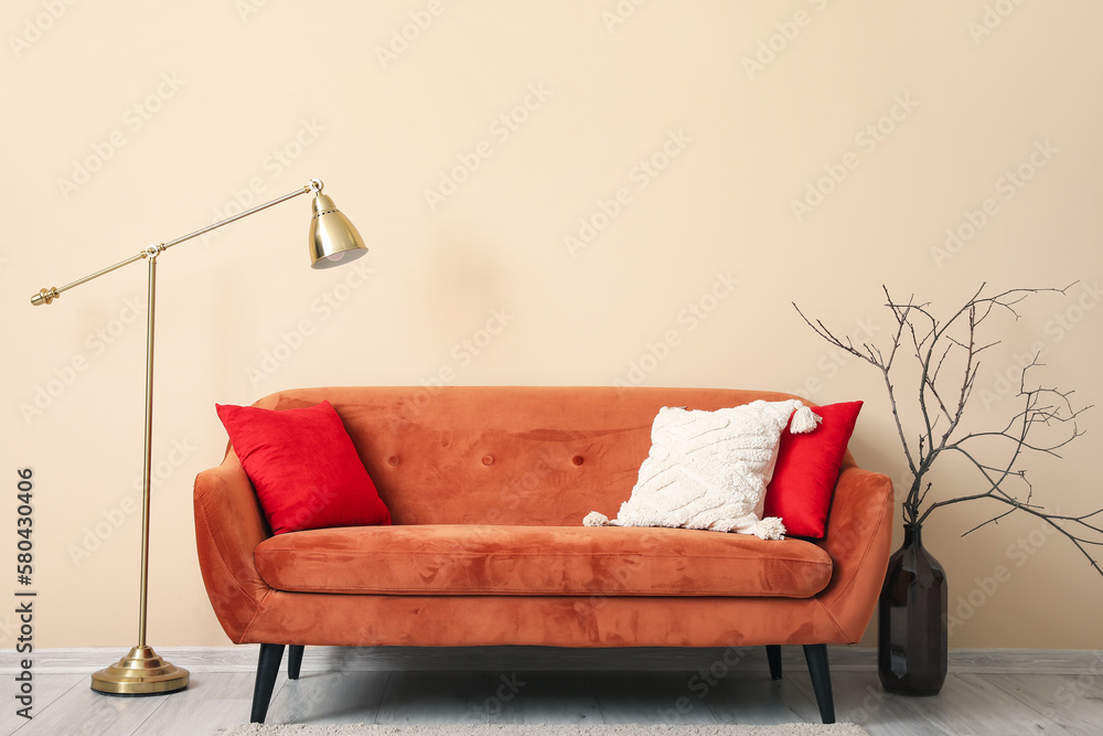 Brown sofa with cushions, lamp and branches in vase near beige wall