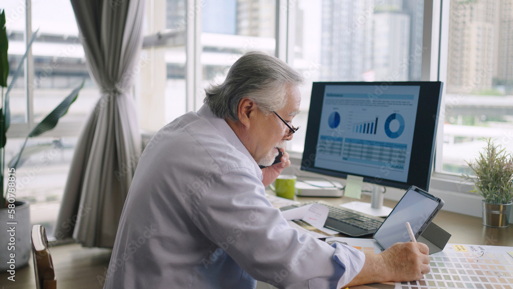 Asian senior mature man working with computers and tablet on table desk at home and talking to custo