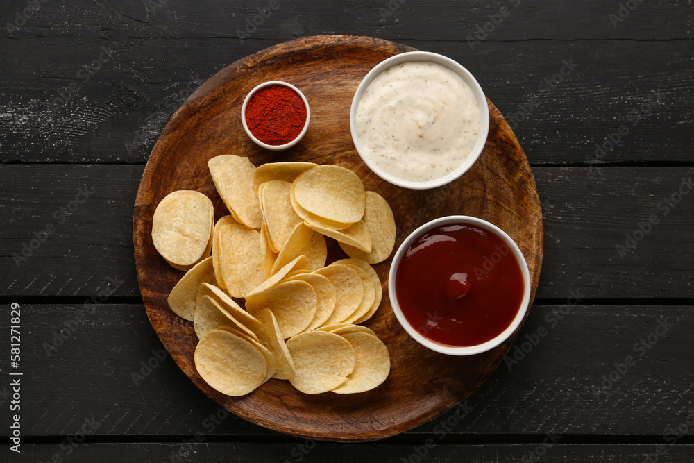 Board with delicious potato chips, ketchup, sauce and paprika on black wooden background