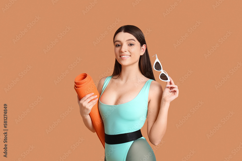 Sporty young woman with yoga mat and sunglasses on beige background