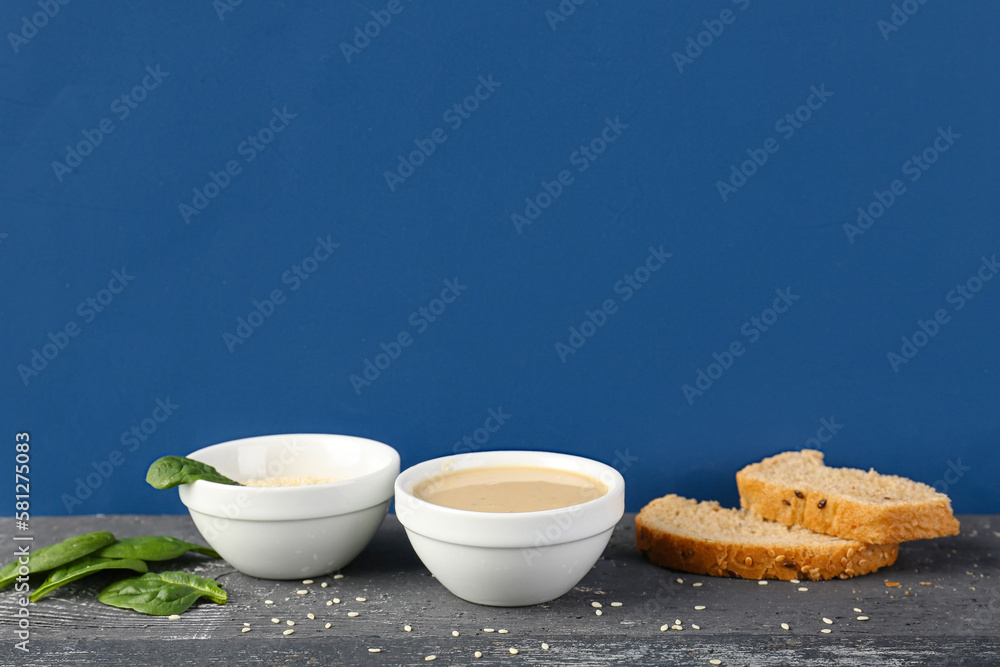 Bowls with tahini, sesame seeds, bread and spinach on dark wooden table near color wall