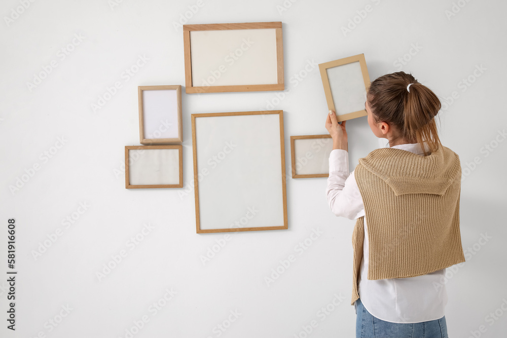 Young woman hanging blank frame on light wall at home, back view