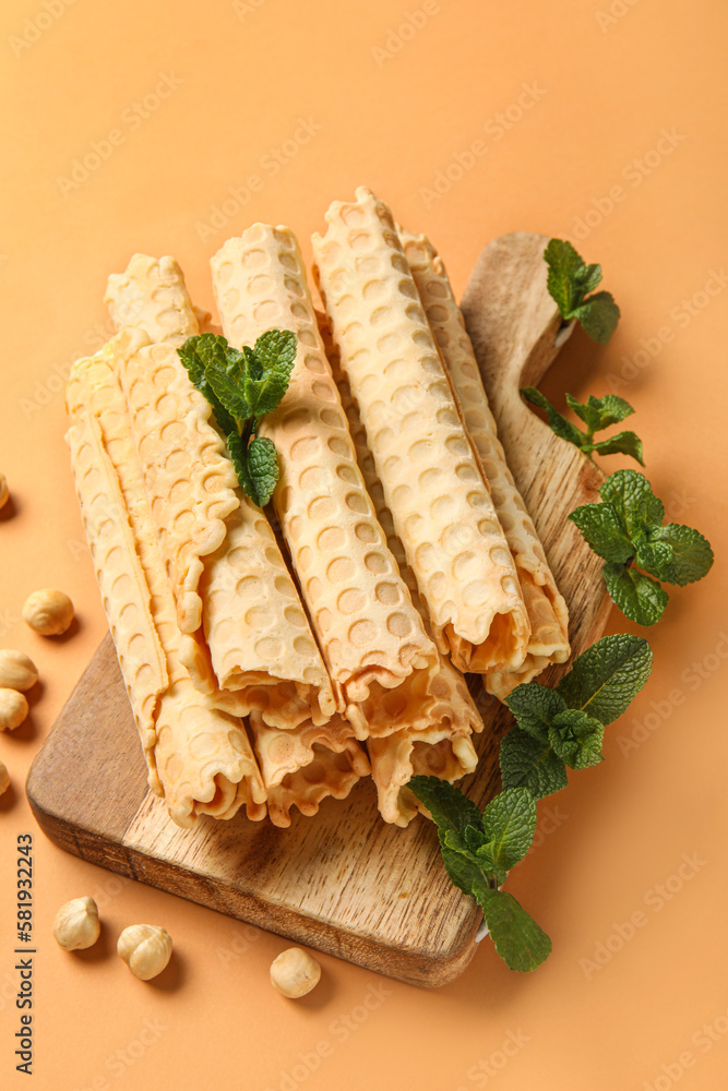 Board with delicious wafer rolls, mint and hazelnuts on orange background