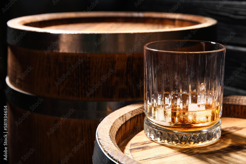 Glass of cold whiskey and oak barrels on dark wooden background