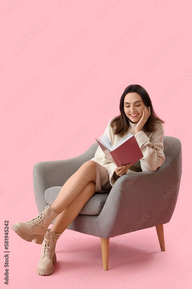 Young woman reading book in grey armchair on pink background