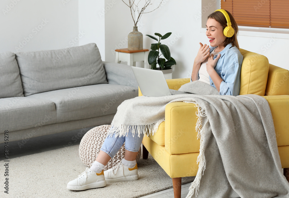 Young woman with laptop watching video in yellow armchair at home