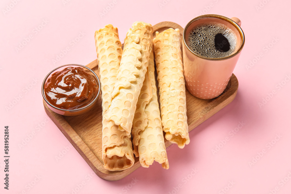 Board with delicious wafer rolls, cup of coffee and boiled condensed milk on pink background