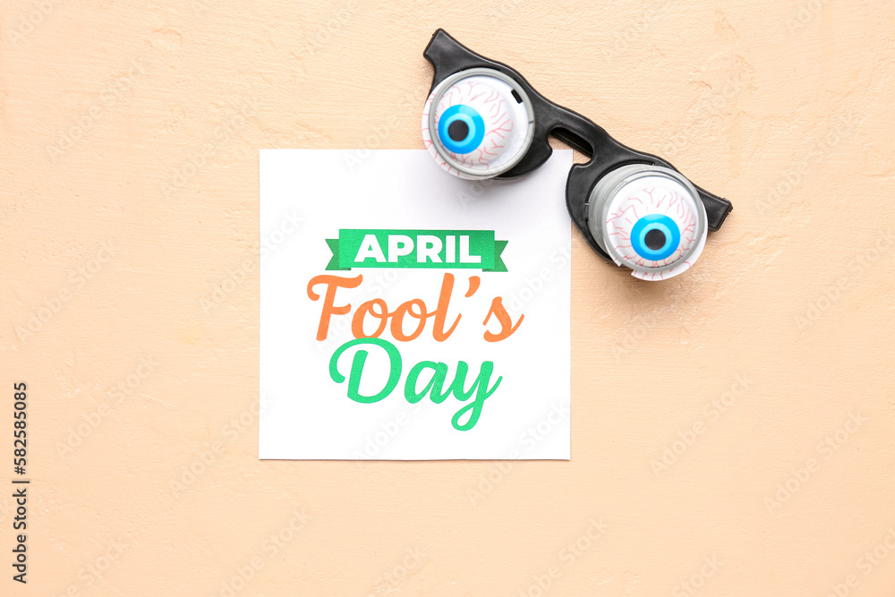 Paper with text APRIL FOOLS DAY and funny eyeglasses on beige background