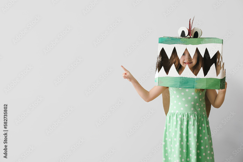 Little girl in cardboard dinosaur costume pointing at something near light wall