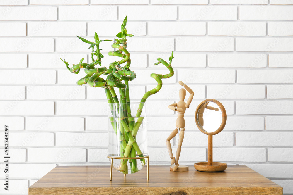 Vase with bamboo plant, wooden mannequin and mirror on table near brick wall