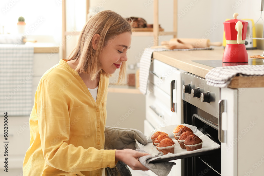 Young woman taking tray with cupcakes from oven in kitchen