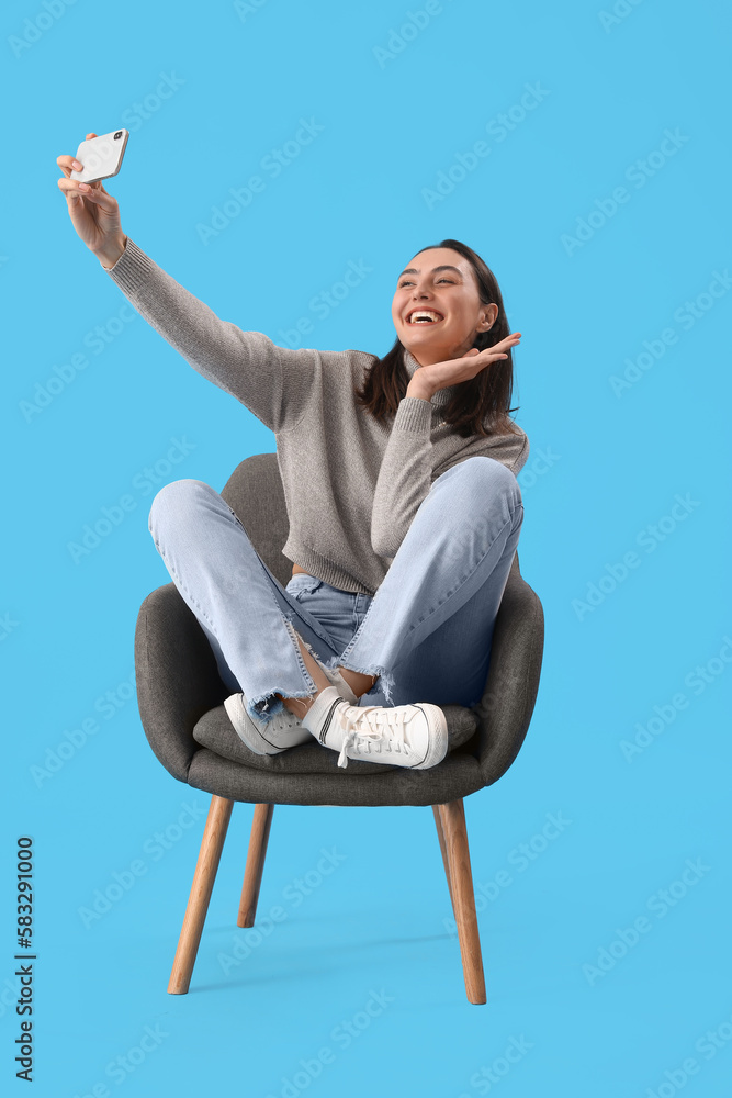 Young woman taking selfie in black armchair on blue background