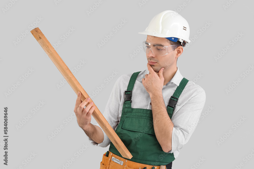 Thoughtful young carpenter with wooden plank on grey background