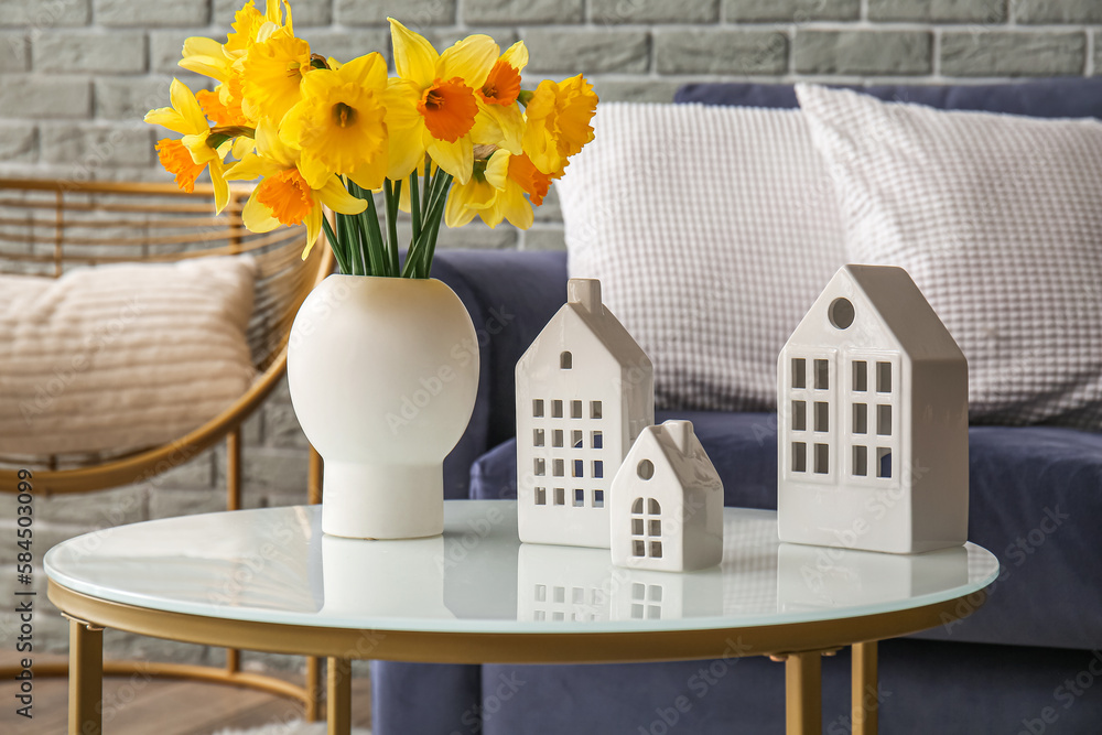 Vase with narcissus flowers and ceramic houses on coffee table in living room