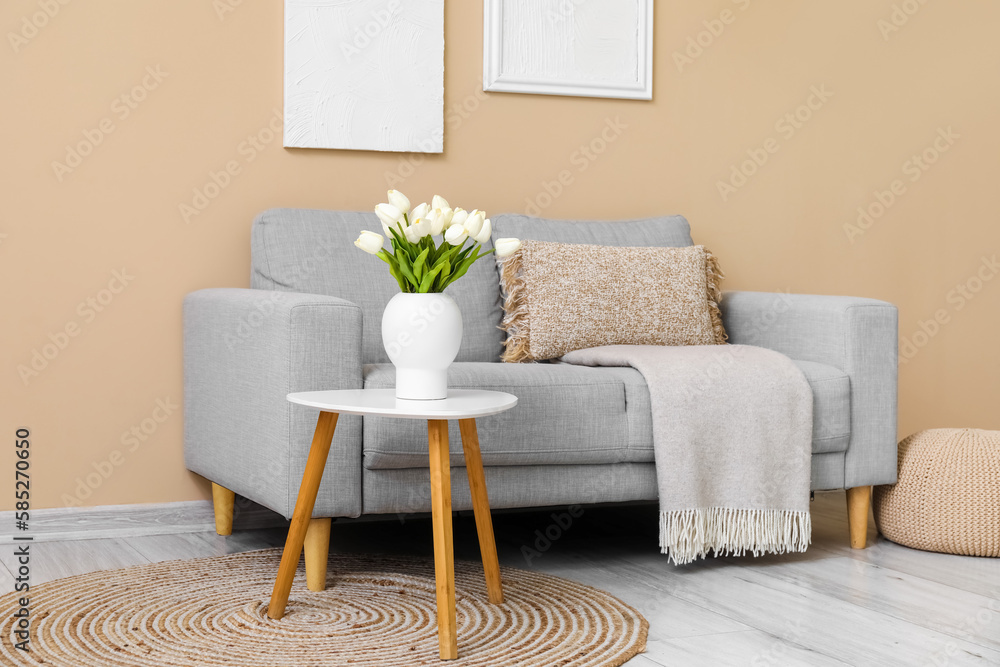 Cozy grey sofa with cushion and vase of tulip flowers on table near beige wall