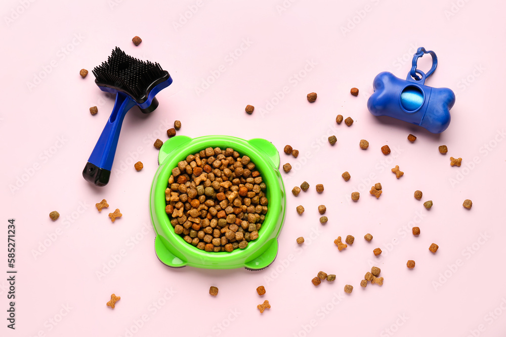 Bowl with dry pet food, grooming brush and waste bags on pink background