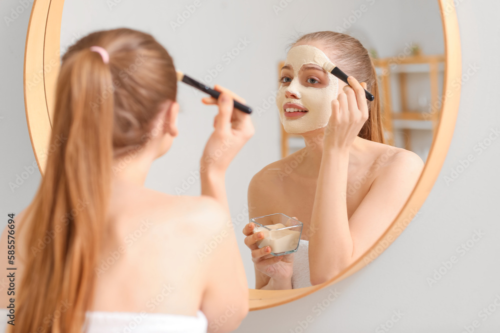 Young woman applying turmeric mask with brush near mirror at home