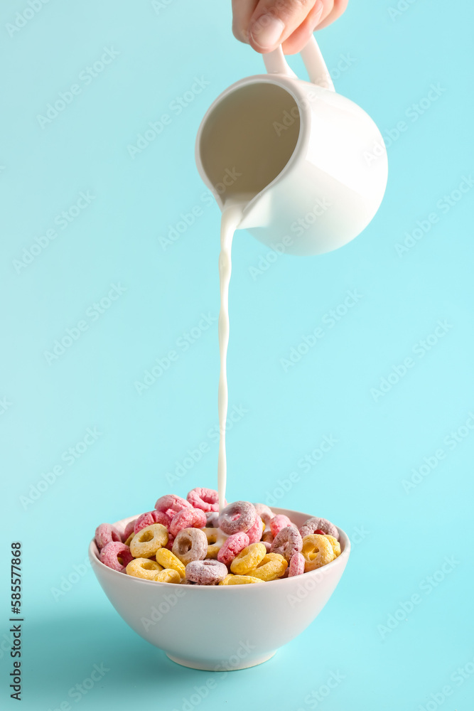 Female hand pouring milk from pitcher into bowl with colorful cereal rings on turquoise background