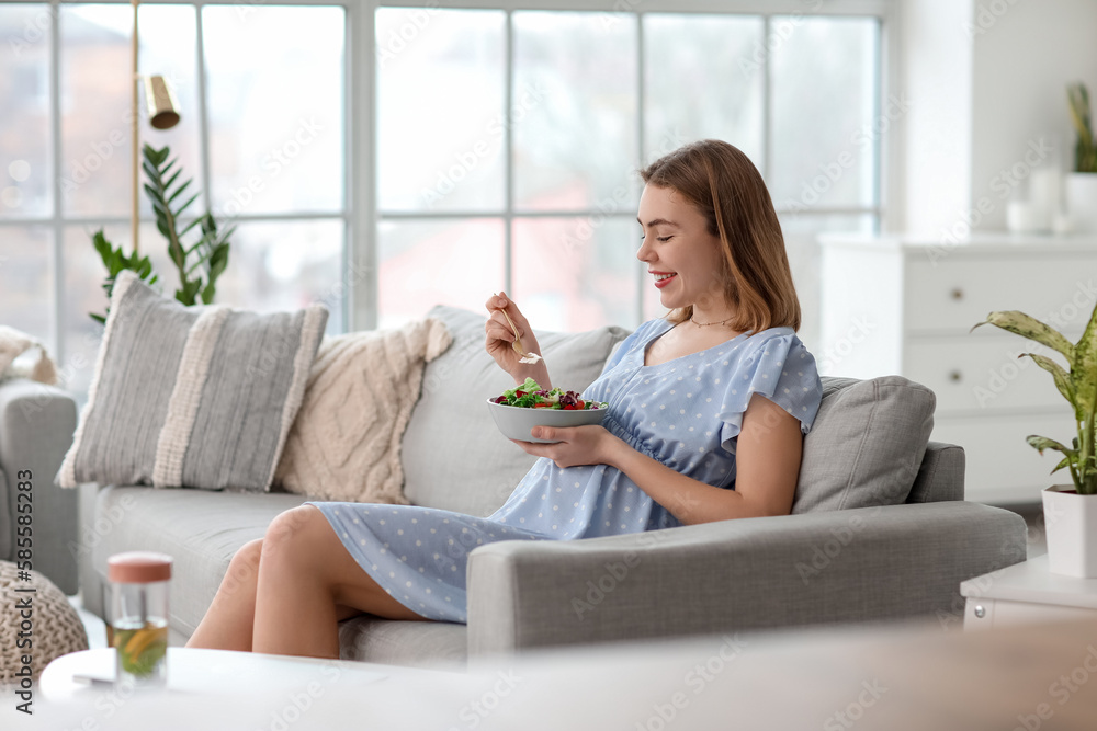 Pregnant young woman eating vegetable salad on sofa at home