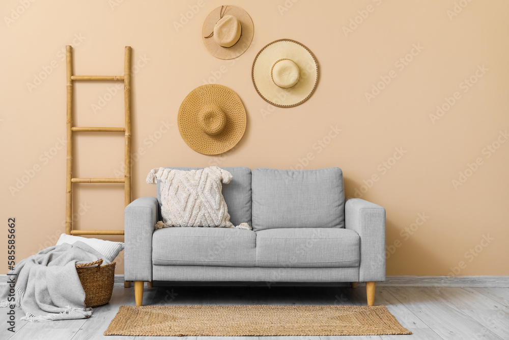 Cozy grey sofa with cushion, plaid in basket, ladder and hanging hats near beige wall