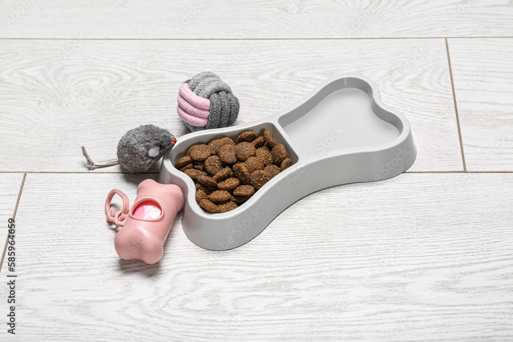 Bowl of dry pet food, waste bags and toys on light wooden background