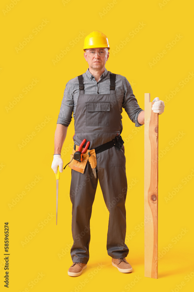 Mature carpenter with hacksaw and wooden plank on yellow background