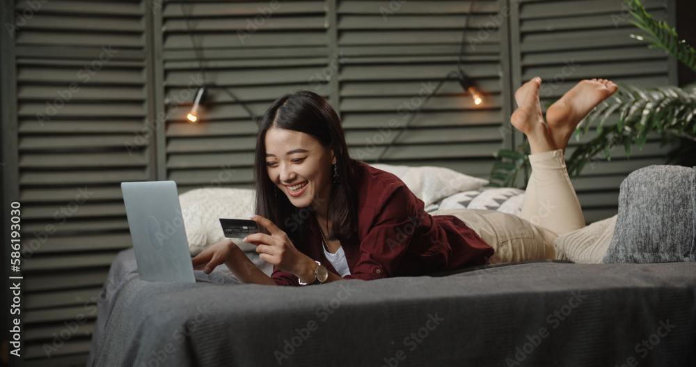 Asian girl lying on large bed with her laptop, having online shopping, keying in the card and happil