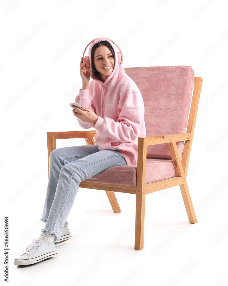 Beautiful woman with headphones and mobile phone sitting in pink armchair on white background