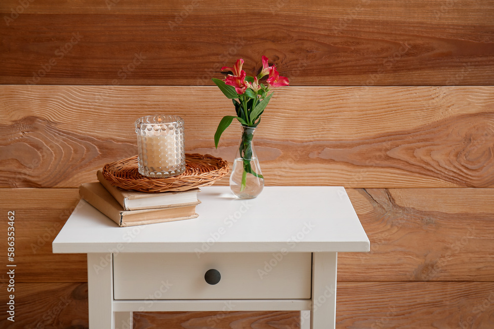 Burning candle, books and vase with flowers on table near wooden wall