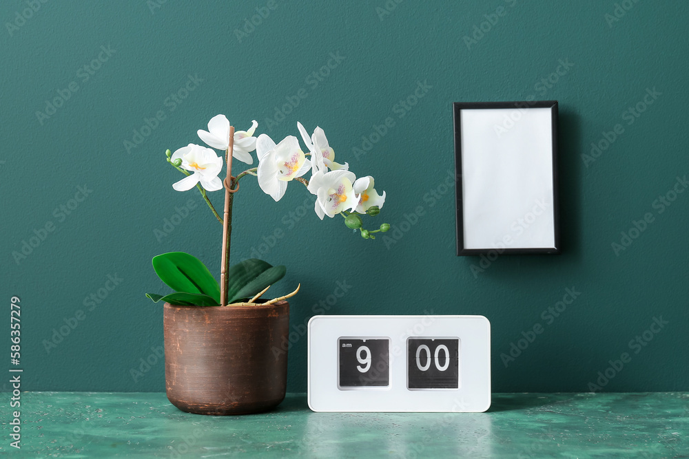 Artificial flower with frame and clock on table near green wall