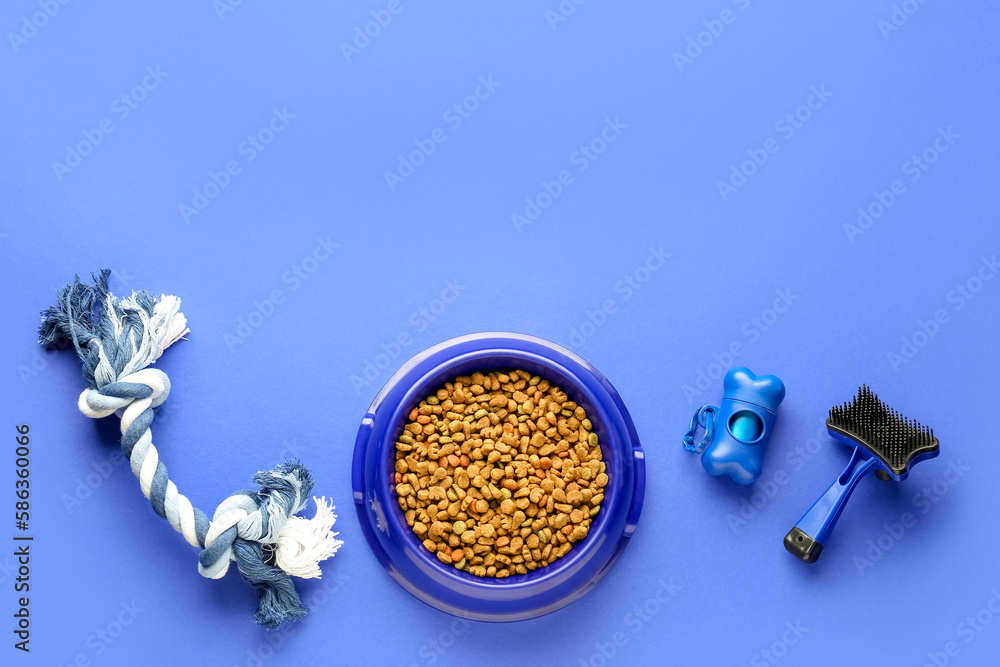 Bowl of dry pet food, toy, waste bags and grooming brush on color background