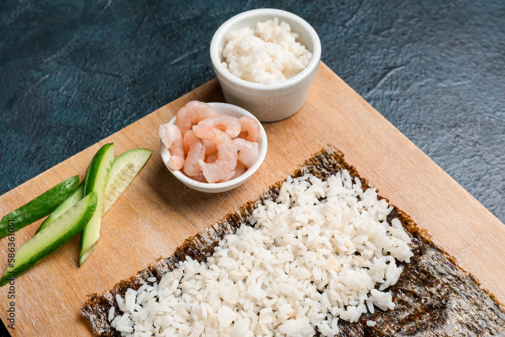 Wooden board with nori and ingredients for preparing sushi rolls on dark background, closeup