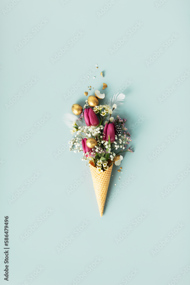 Ice cream cone with beautiful flowers and golden easter eggs on blue background flat lay