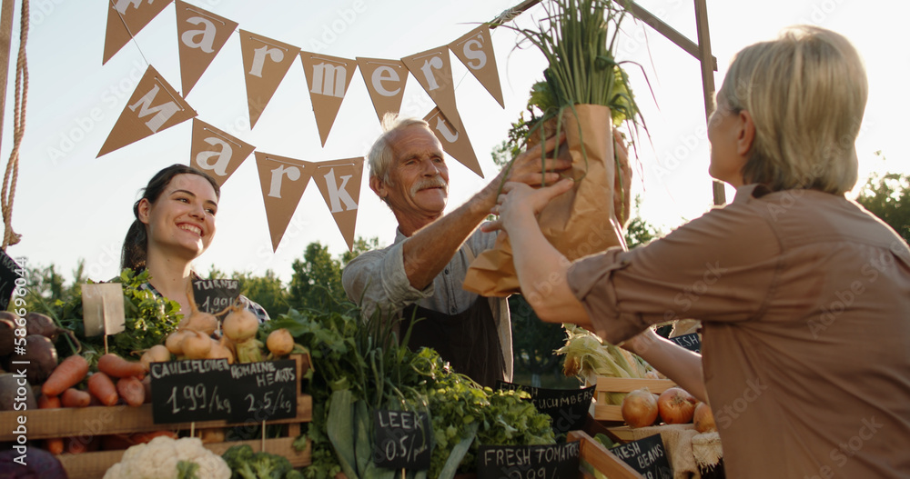 Senior woman buying groceries at local farmers market. Senior man and his daughter offering fresh fr