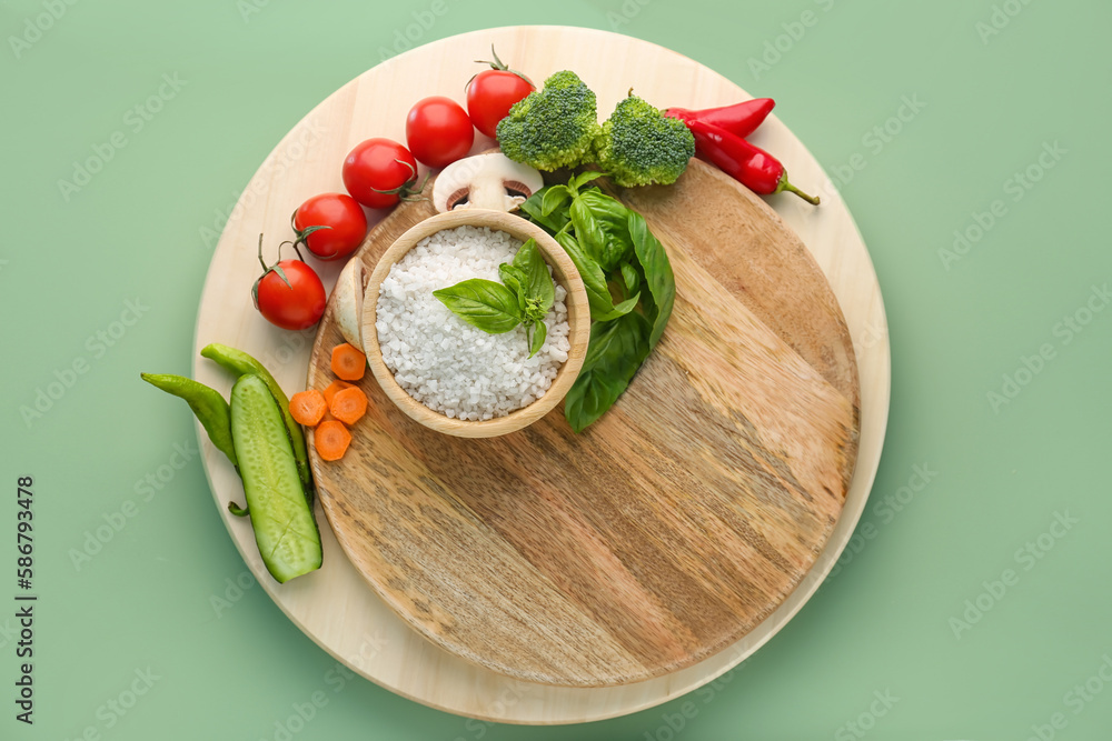 Wooden cutting board with bowl of sea salt and fresh vegetables on color background