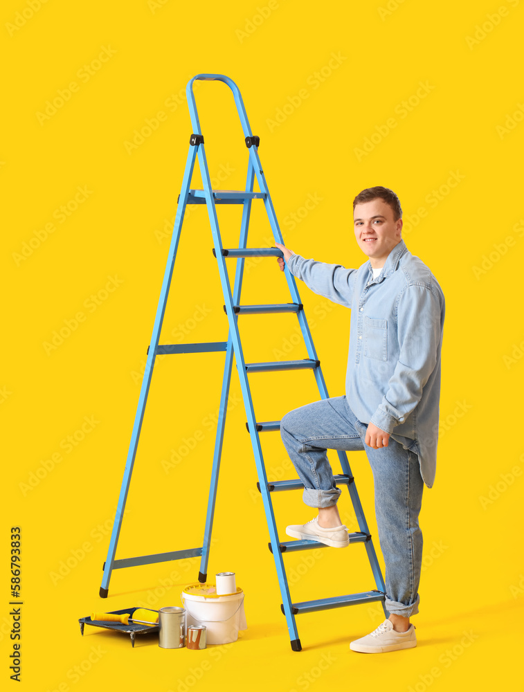 Young man with ladder and cans of paint on yellow background