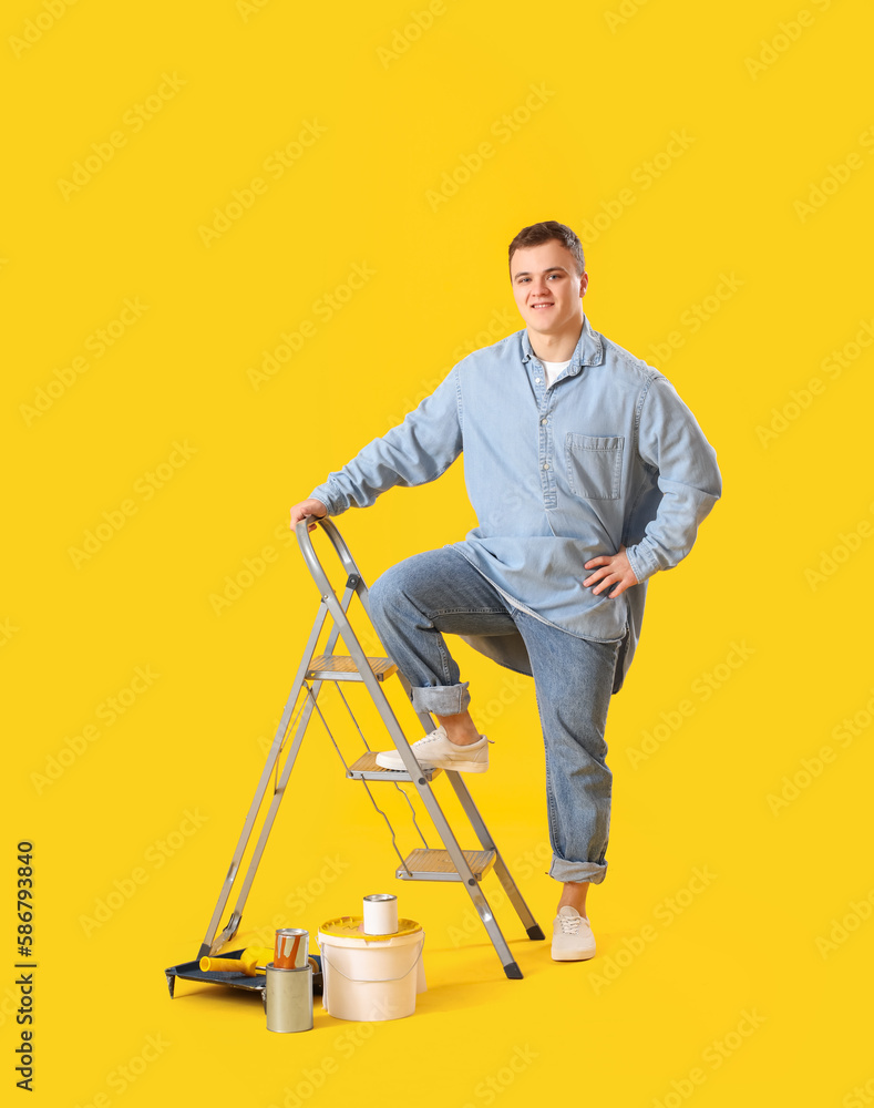 Young man with ladder and cans of paint on yellow background